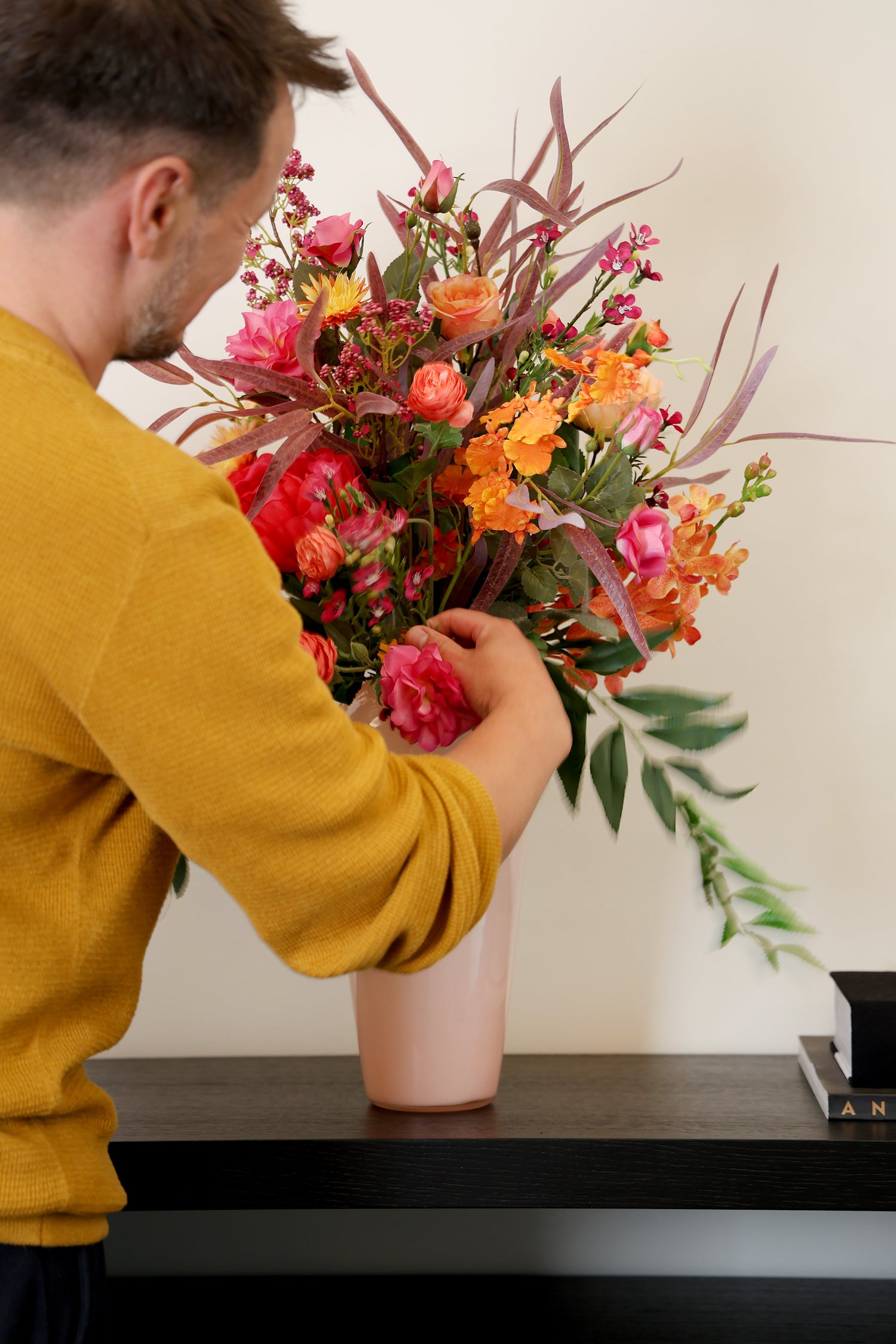 Künstlicher Blumenstrauß Flower Cocktail von Lumisa’s Choice in kräftigen Farben in  apricot farbener Vase auf schwarzem Sideboard wird von Person arrangiert.