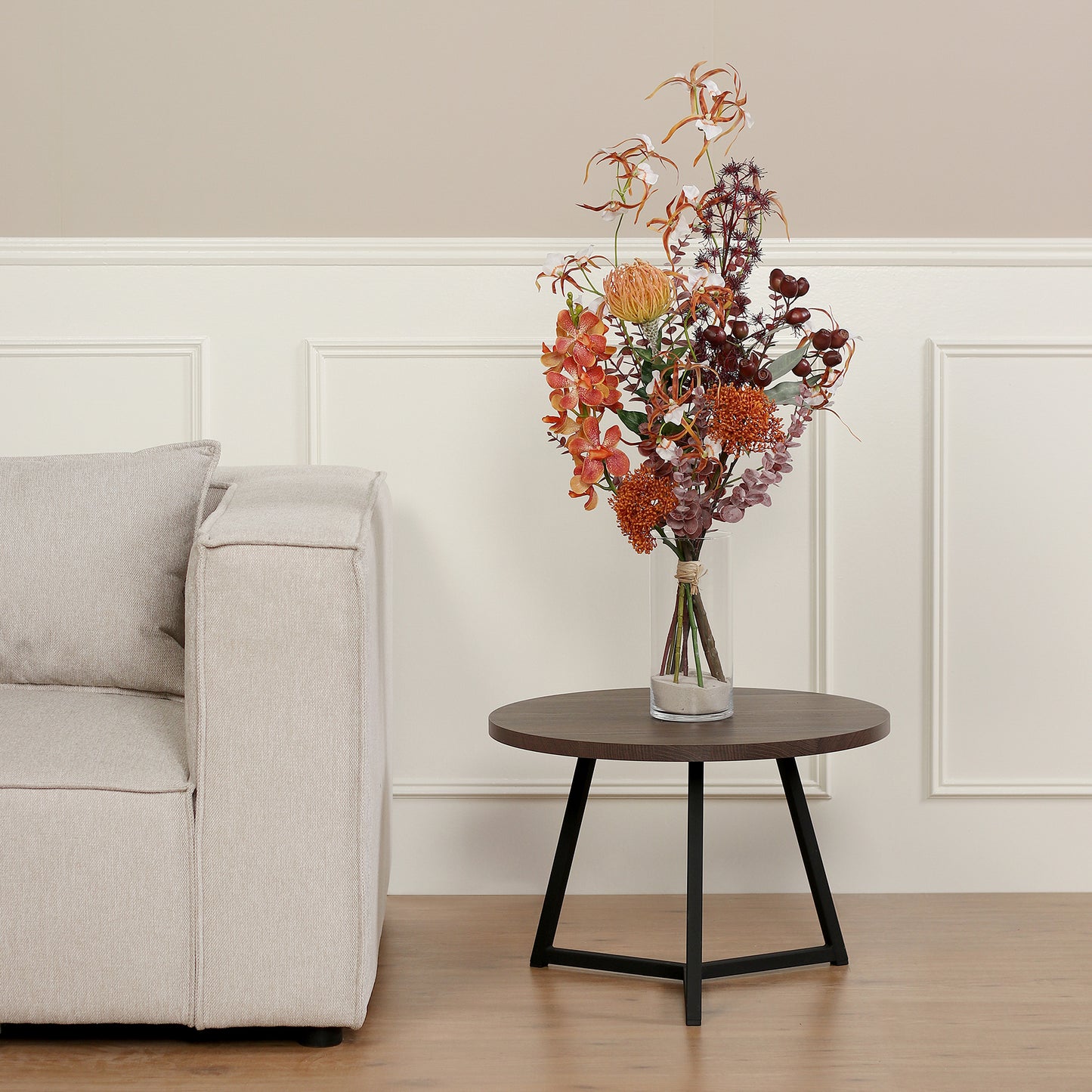 Künstlicher Blumenstrauß African Sunset von Lumisa’s Choice in warmen Farben in Glasvase als stilvolle Dekoration neben hellem Sofa.