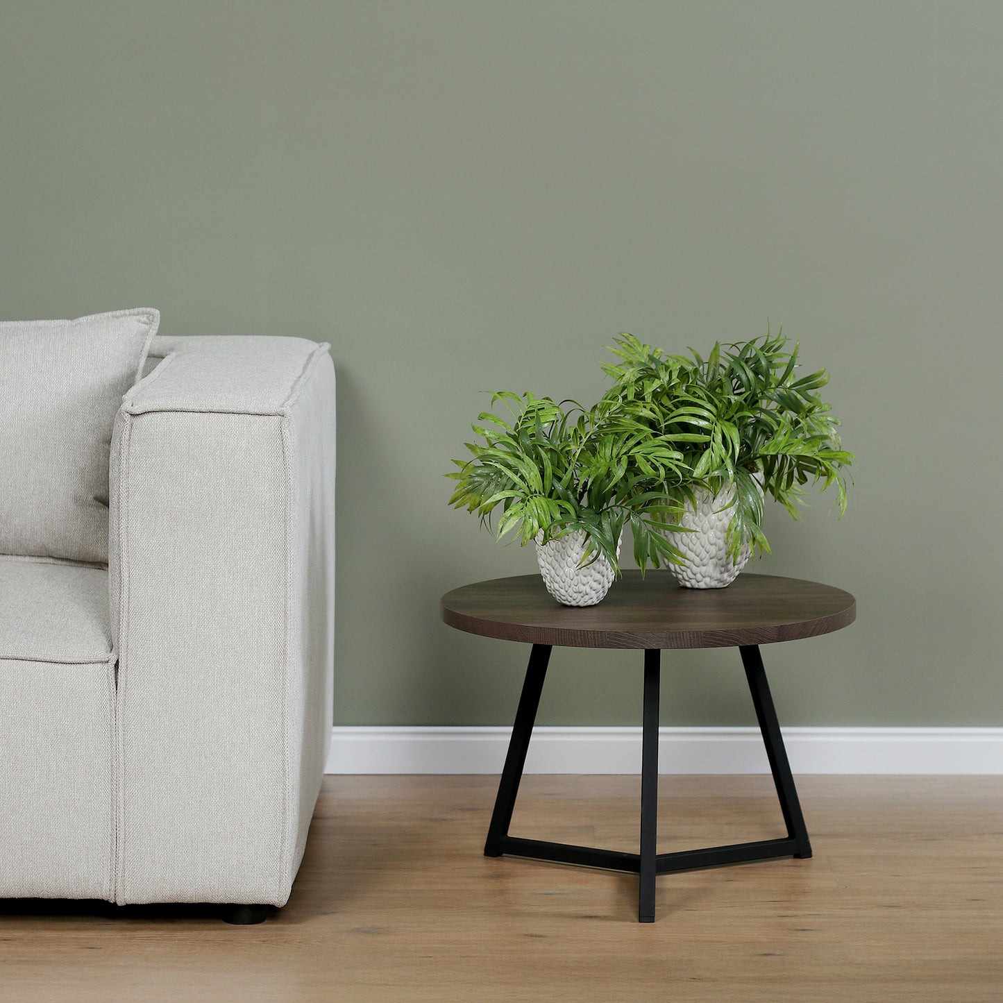 Zwei künstliche Chico-Pflanzen im Set in weißen Keramiktöpfen Lumisa’s Choice auf rundem Holztisch neben hellem Sofa.