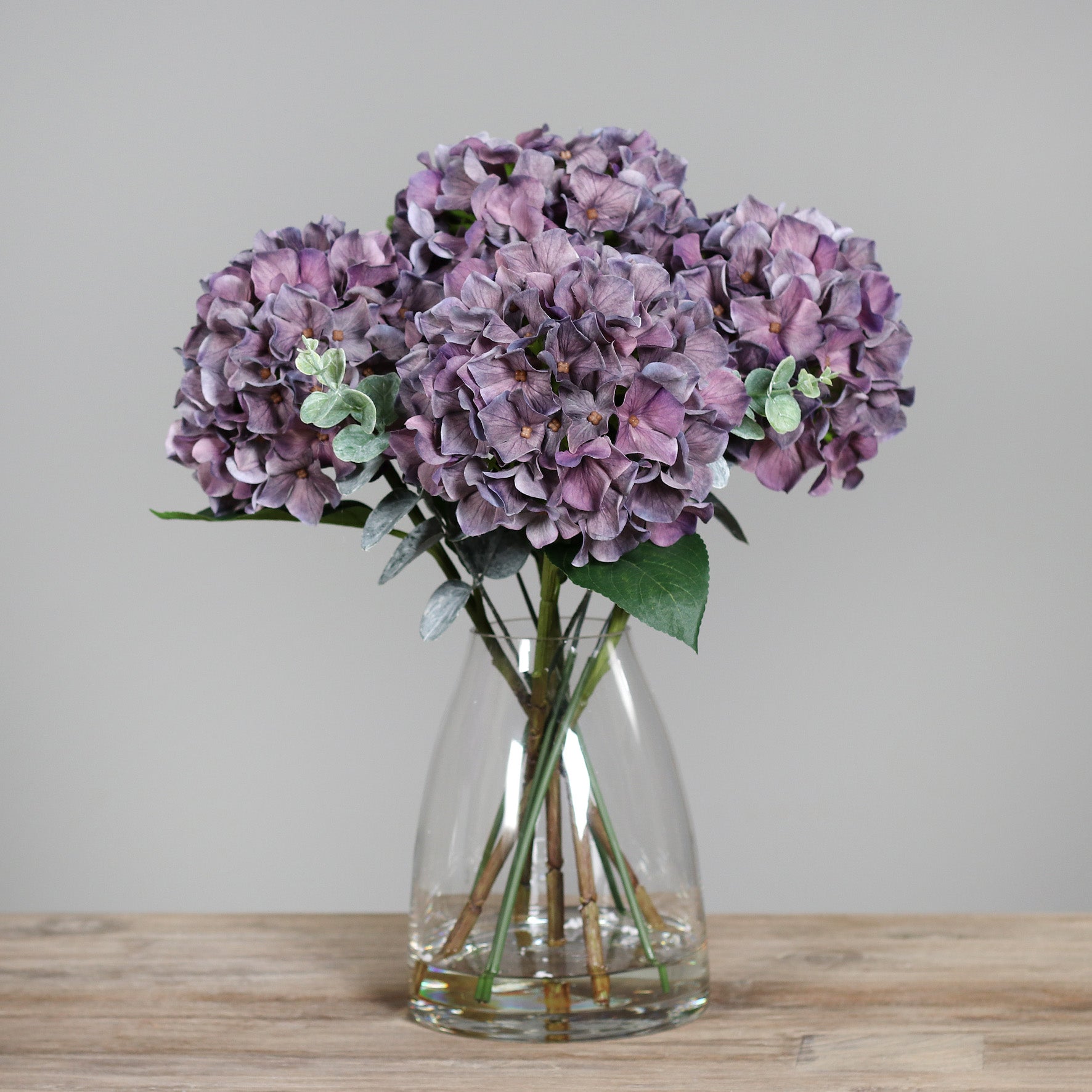 Künstliche mauve farbene Hortensie in Glasvase auf Holztisch