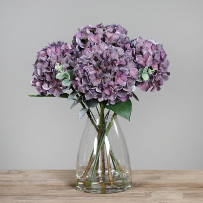Künstliche mauve farbene Hortensie in Glasvase auf Holztisch